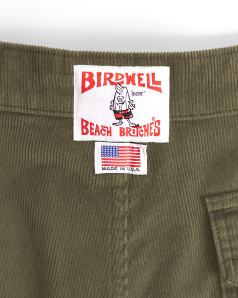 Close-up of a Birdwell Beach Britches clothing label on olive green fabric, featuring a cartoon bird, company logo, and a small American flag tag that says MADE IN U.S.A. This label is sewn onto their Classic Corduroy Shorts - Fatigue, which feature a classic retro fit and an adjustable drawcord waist.
