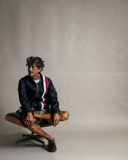 A man with curly hair sits cross-legged on a chair, wearing the Racing Jacket 26 - Black/White/Red, tan shorts, and dark shoes, against a plain beige background.