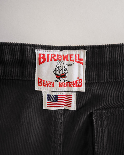Close-up of charcoal Classic Corduroy Pants in 100% cotton corduroy, featuring a stitched Birdwell Beach Britches brand label with a cartoon mascot positioned above a small American flag patch.