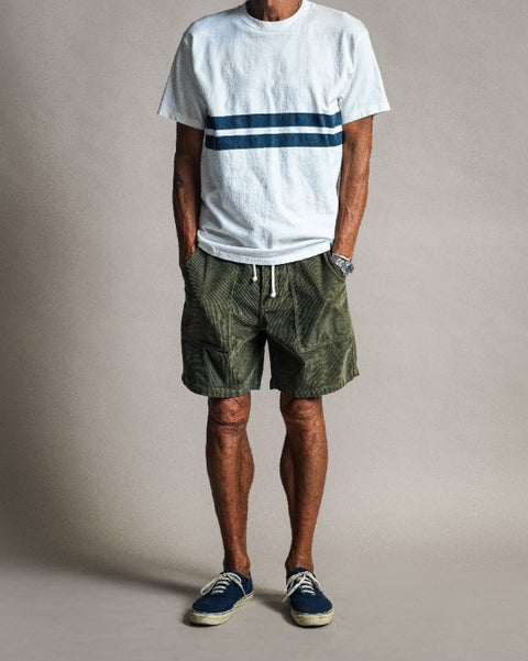 A person wearing a Comp Stripe T-Shirt - Vintage White, olive green shorts with a drawstring, and blue canvas sneakers.