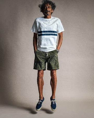A man captured mid-air wearing a Comp Stripe T-Shirt - Vintage White and olive green patterned shorts.