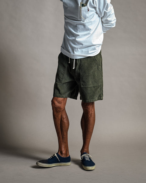 A person wearing a white long-sleeved shirt and olive green Classic Corduroy Shorts - Sand with a drawstring waist, paired with dark blue sneakers.