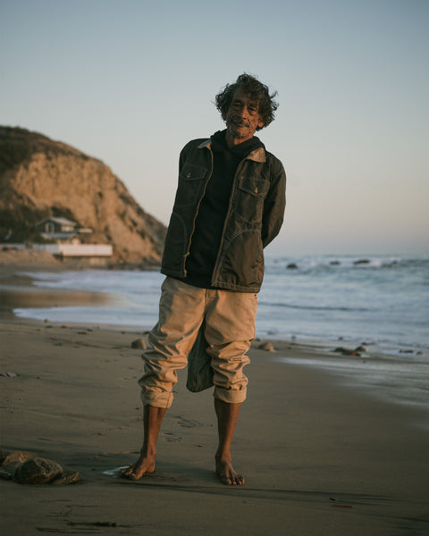 A man wearing a dark Capistrano Jacket - Frogskin/Acorn and tan pants standing barefoot on a sandy beach.
