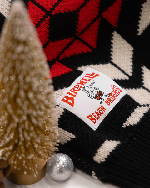Close up detail shot of the license plate label stitched onto the sweater. Gold miniature christmas tree and silver ball ornament in the foreground.