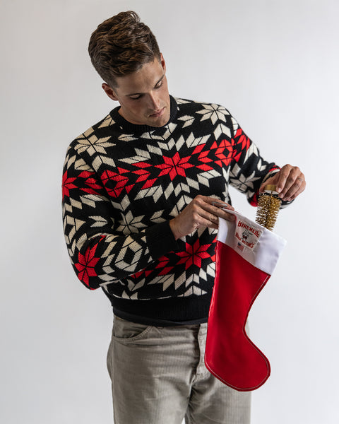Model in front of white background wearing the Christmas Sweater while holding the Birdwell SurfNyl™ stocking while putting a gold miniature tree in the stocking. 