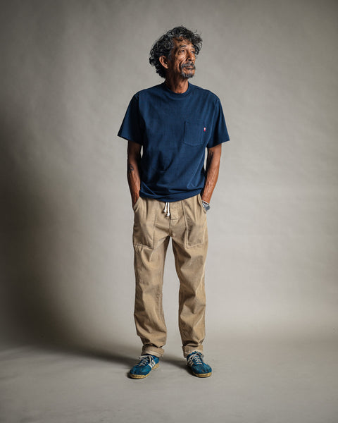 A man with curly hair and a beard stands in front of a neutral background, wearing the High Tee Pocket - Navy, paired with beige drawstring pants and blue sneakers. The navy t-shirt is made in the USA from 100% cotton for extra comfort. He has his hands in his pockets and gazes slightly to the side.