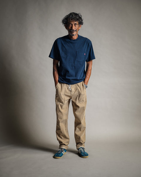 A man with curly hair and a beard stands smiling, wearing the High Tee Pocket - Navy, 100% cotton t-shirt, along with beige drawstring pants and blue sneakers, against a neutral studio background.