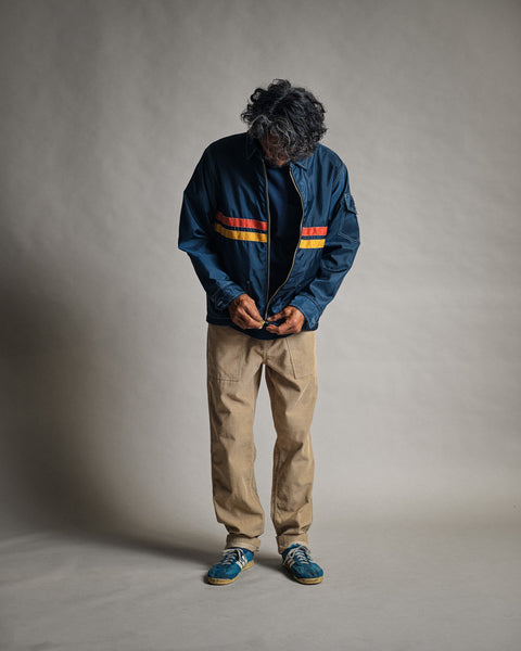 A person with dark, curly hair looks down while buttoning a Comp Jacket 26 in Navy/Gold/Paprika. They wear tan pants and blue shoes, standing against a plain, light gray background.