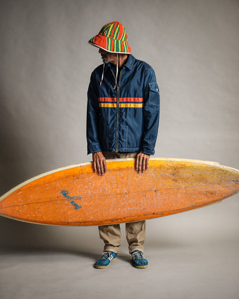 A person wearing a colorful striped hat, tan pants, sneakers, and the Comp Jacket 26 in Navy/Gold/Paprika stands holding a large, worn orange surfboard made from American-made SurfNyl against a plain background.