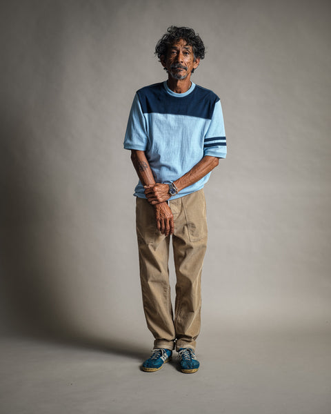 A man with curly hair and a mustache stands against a plain background, wearing the Special Teez Champ in Arctic Blue/Navy—crafted from premium USA-grown cotton—paired with beige pants and blue sneakers, his hands clasped in front of him.