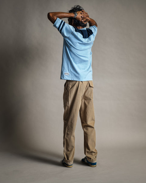 A person stands facing away, hands resting on their head. They wear the Special Teez Champ in Arctic Blue/Navy—a premium t-shirt made from USA-grown cotton—paired with tan pants and striped shoes, against a neutral background.