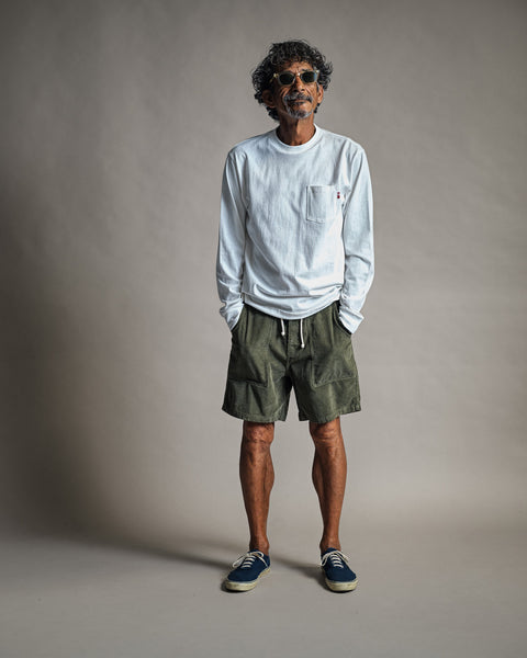 A man with curly hair and sunglasses stands against a gray backdrop, smiling with his hands in his pockets while wearing the High Tee Pocket Long Sleeve in Vintage White, paired with olive green drawstring shorts and navy sneakers.