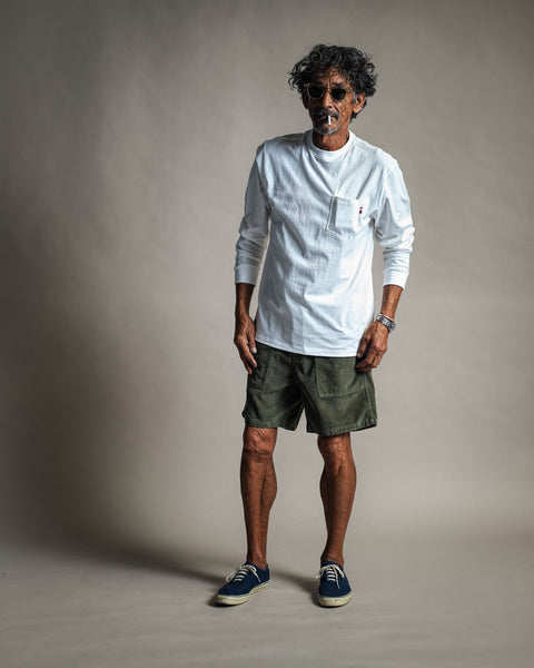 A man with curly hair and sunglasses wears the High Tee Pocket Long Sleeve in Vintage White, paired with green shorts and blue sneakers. He stands against a plain gray background, looking relaxed with his hands by his sides.