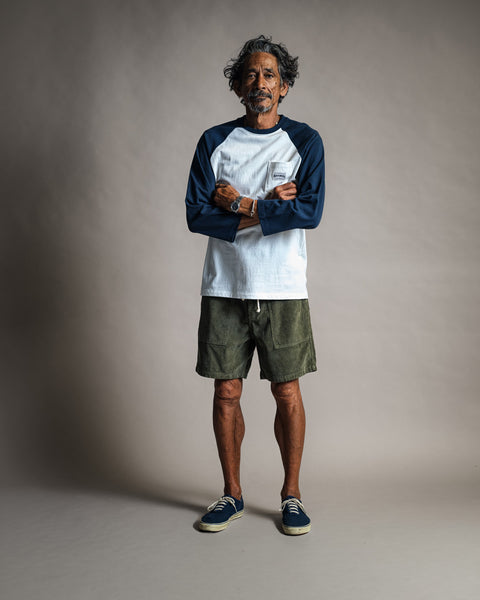 A smiling man with curly hair stands with his arms crossed, wearing the Special Teez Pastime t-shirt in vintage white and navy. He pairs it with olive green shorts and navy sneakers, against a plain gray background.