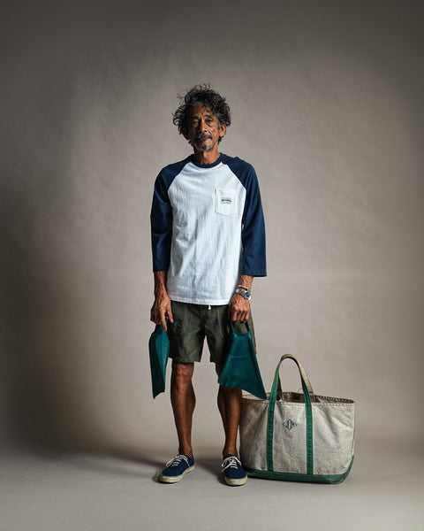 A man with curly hair stands against a plain background wearing the Special Teez Pastime vintage white/navy 3/4 sleeve raglan t-shirt, shorts, and sneakers. He holds two green pouches, with a large beige and green tote bag placed at his feet.