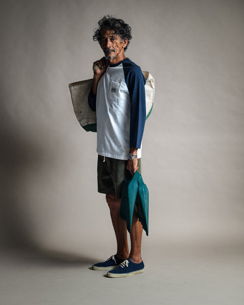 A man stands in a studio holding a green jacket and a large bag over his shoulder. He wears the Special Teez Pastime raglan t-shirt in Vintage White/Navy—crafted from USA-grown cotton—paired with green shorts and blue sneakers, looking confidently at the camera.