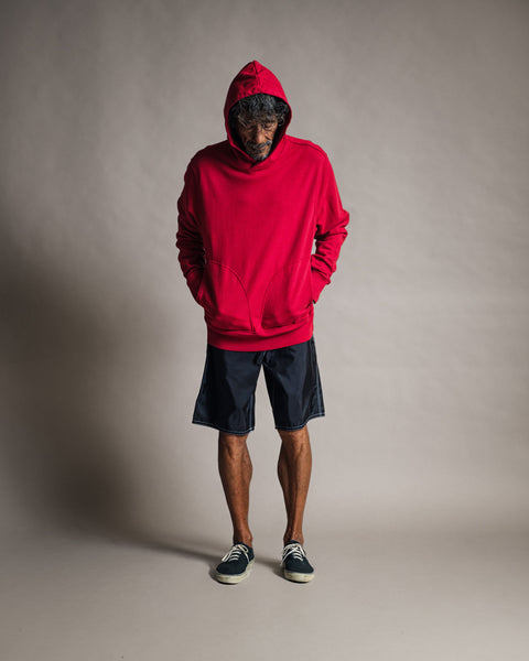 A person stands facing downward, hands tucked into the pocket of the Harmony Hoodie - Red, a premium piece crafted from soft French terry in a vibrant red hue. The hoodie is paired with dark shorts and black sneakers, set against a plain, light backdrop.