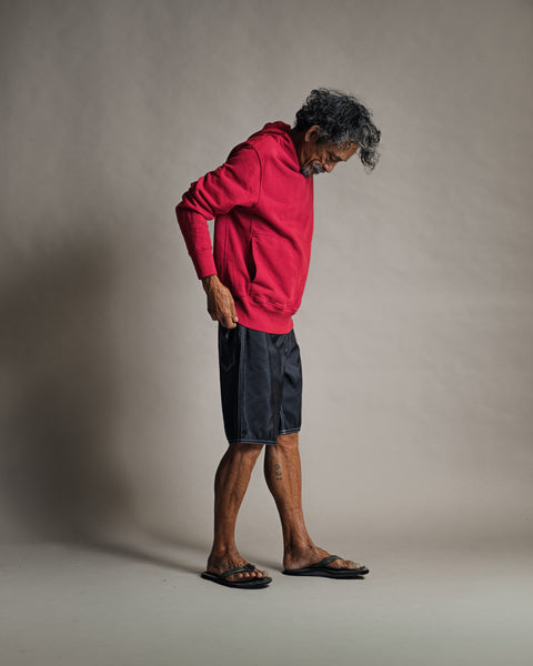 An older man with curly gray hair stands sideways against a plain light background, looking down while wearing the Harmony Hoodie - Red, paired with black shorts and sandals.