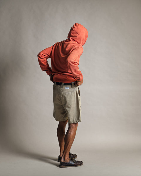 A person wearing the Harmony Hoodie - Paprika, crafted from 100% cotton French terry, along with khaki shorts and brown shoes, stands with hands on hips facing away from the camera against a plain, light-colored background.