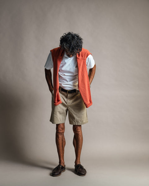 A person with curly hair stands, looking down, wearing a white t-shirt, tan shorts, brown shoes, and the Harmony Hoodie - Paprika—a premium 100% cotton French terry sweatshirt in a vibrant orange shade—draped over their shoulders against a plain background.