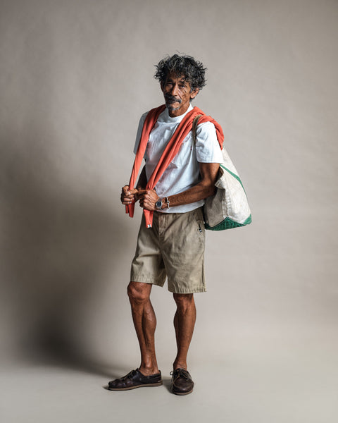 An older man with gray curly hair stands in shorts and a white t-shirt, wearing the Harmony Hoodie in Paprika draped over his shoulders and carrying a large bag. He stands against a plain, neutral background.