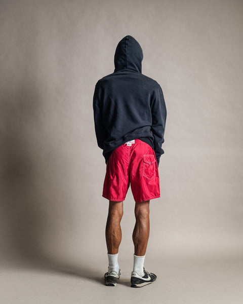 A person stands facing away, wearing the Harmony Hoodie in navy with the hood up, paired with bright red shorts, white socks, and sneakers. The hoodie is crafted from soft French terry and made in the USA. The background is plain and neutral.