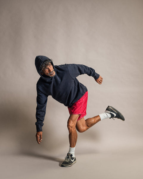 An older man wearing the Harmony Hoodie - Navy, made in the USA, and red shorts balances on one leg with his body tilted and arms outstretched against a plain gray background.