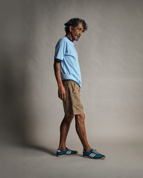 An older man with curly gray hair stands sideways, smiling. He wears the Special Teez Ringer in Arctic Blue/Navy—a relaxed fit, premium t-shirt made from USA-grown cotton—paired with tan shorts and blue Adidas sneakers against a neutral studio background.