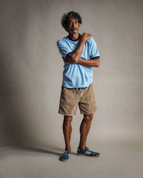 A man with curly hair stands against a neutral background, wearing the Special Teez Ringer in Arctic Blue/Navy—crafted from premium USA-grown cotton—paired with tan shorts and blue shoes. He crosses one arm over his body, touching his opposite shoulder, and gazes slightly to the side.