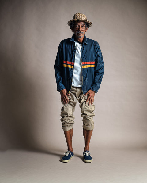 An older man stands against a plain backdrop, wearing a woven hat, the Comp Jacket 26 in Navy/Gold/Paprika (American-made) with orange stripes, a white t-shirt, rolled-up khaki pants, and navy sneakers. He looks relaxed, with his hands resting on his knees.