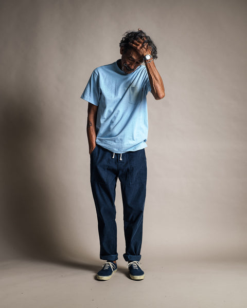 A man stands against a plain background, wearing the High Tee Pocket in Arctic Blue—a light blue 100% cotton pocket t-shirt—paired with dark blue pants and blue sneakers. He is looking down and scratching his head with one hand.