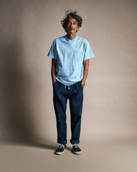 A man with curly hair and a beard smiles, standing against a plain background. He wears the High Tee Pocket in Arctic Blue—made from 100% cotton—along with navy drawstring pants and navy sneakers. His hands are in his pockets.