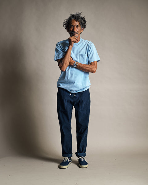 A man with curly hair stands in front of a plain background, wearing the High Tee Pocket in Arctic Blue, paired with dark blue pants and blue sneakers. He holds his chin with one hand, looking thoughtful.