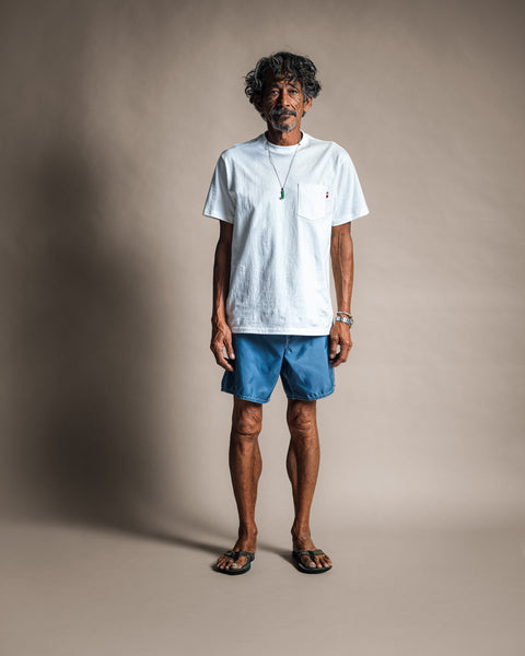 A man with curly hair stands on a plain background wearing the High Tee Pocket in Vintage White, blue shorts, sandals, a green necklace, and bracelets. He looks directly at the camera with a neutral expression.