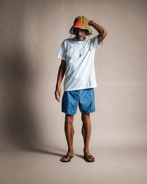 A man stands in front of a plain background wearing a colorful striped hat, blue shorts, flip-flops, and a beaded necklace, with one hand touching his hat. He completes his look with the High Tee Pocket in Vintage White—a premium pocket t-shirt crafted from 100% cotton and proudly made in the USA.