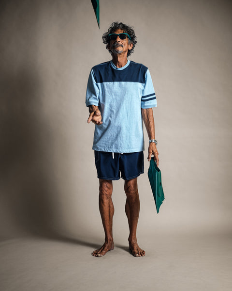 A barefoot man in sunglasses wears the Special Teez Champ shirt in Arctic Blue/Navy—a relaxed-fit, USA-grown cotton color-block top—and navy shorts as he tosses a green cap into the air while holding another cap in his left hand, standing against a plain beige backdrop.