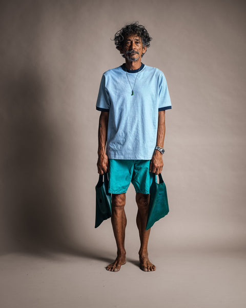 A barefoot man with curly hair wears the Special Teez Ringer in Arctic Blue/Navy—crafted from USA-grown cotton—paired with teal shorts and holding green swim fins in each hand, standing in front of a plain, neutral backdrop.