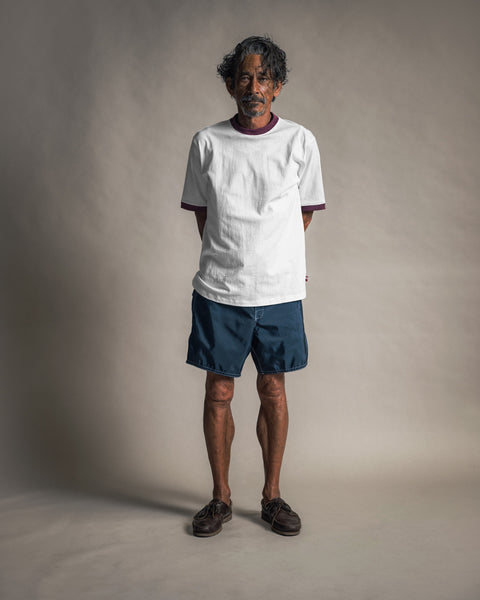 A man with curly hair and a mustache stands facing forward, hands behind his back, wearing the Special Teez Ringer in Vintage White/Burgundy, crafted from 100% USA-grown cotton, paired with navy shorts and brown shoes against a plain gray background.