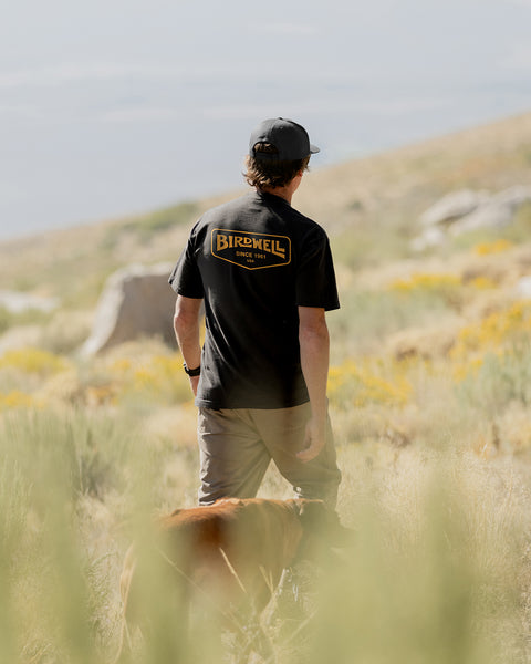 Person wearing he Service T-Shirt in Black with Charcoal Grey Snapback while standing in a field with dog following.