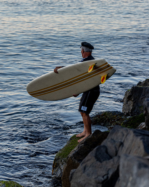 Brian Bent holding a surfboard with three vertical stripes and two yellow fins while wearing the 310 Bent Boardshorts in Black.