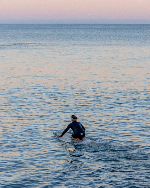 Brian Bent paddling out into the ocean while wearing the 310 Bent in black and a Black wetsuit top. Blue ocean water with a reflection of pink from the sunset.