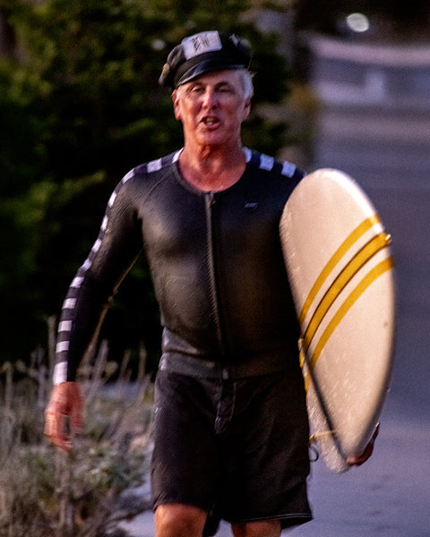 Brian Bent wearing a black wetsuit top with white stripes down the arms, walking forward while holding a board under his left arm. Wearing the 310 Bent in back.