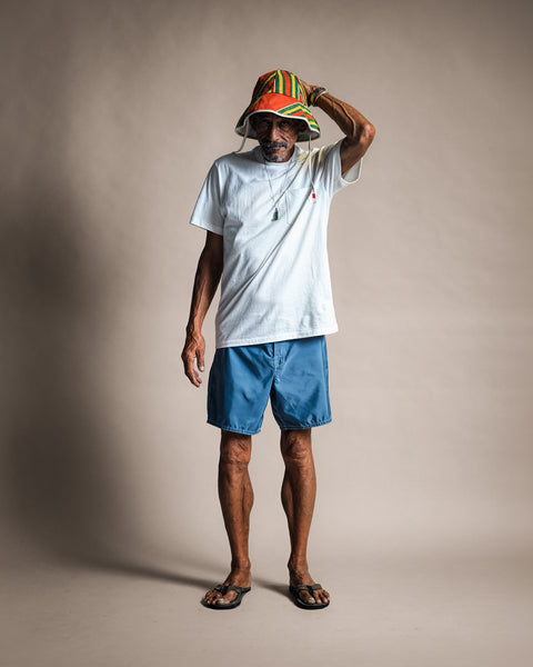 A man wears a High Tee Pocket - Vintage White, blue shorts, flip-flops, and a colorful bucket hat while holding the brim of the hat.