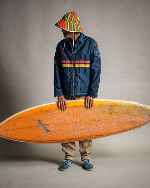A person wearing a Comp Jacket 26 - Navy/Gold/Paprika and a colorful striped bucket hat, holding an orange surfboard.