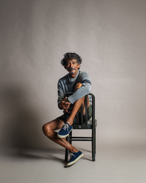 A man wearing a gray and navy Comp Stripe Sweater with navy stripes on the left sleeve, sitting on a wooden chair.