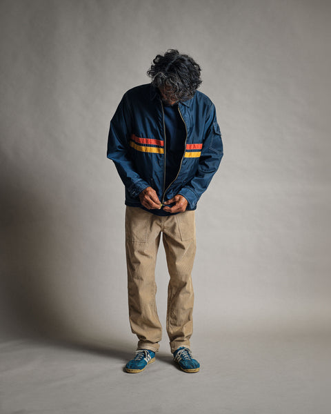 A person zipping up a navy, gold, and paprika Comp Jacket 26 with horizontal stripes across the chest, wearing tan pants and blue sneakers.