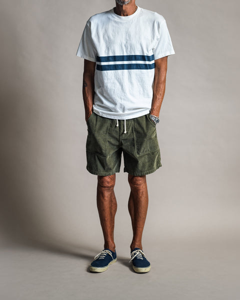 A man wears a Comp Stripe T-Shirt - Vintage White featuring two horizontal blue stripes, olive green shorts with a drawstring waist, and blue sneakers.