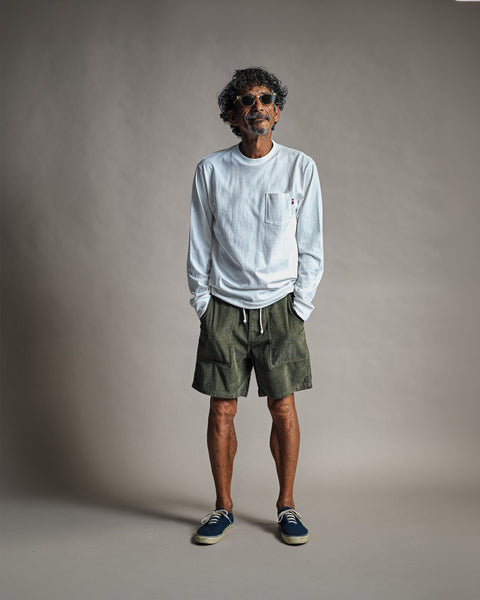A man wearing a white long-sleeve t-shirt, Classic Corduroy Shorts - Olive with an adjustable drawcord waist, and blue sneakers.