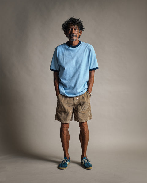 A man stands with his hands in his pockets wearing a Special Teez Ringer - Arctic Blue/Navy, a light blue relaxed fit t-shirt with navy trim, paired with tan shorts.