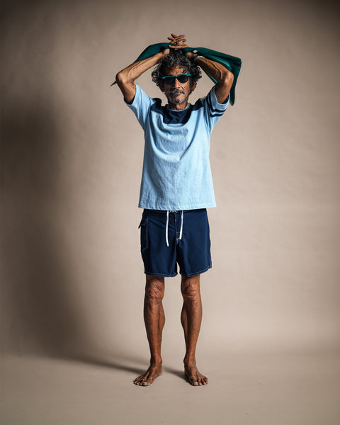 A man wearing sunglasses, a Special Teez Champ - Arctic Blue/Navy t-shirt, and navy board shorts, standing with his hands clasped behind his head.